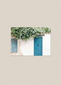 Poziomy plakat przedstawiający biały budynek z turkusowymi drzwiami oraz kwiatami zwisającymi z dachu.