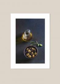 Pionowy plakat przedstawiający oliwki w miseczce  i oliwę w szklanej karafce na ciemnym tle.